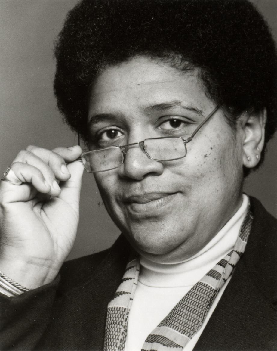 Audre Lorde, female presenting black woman with an afro, holding glasses and looking into the camera