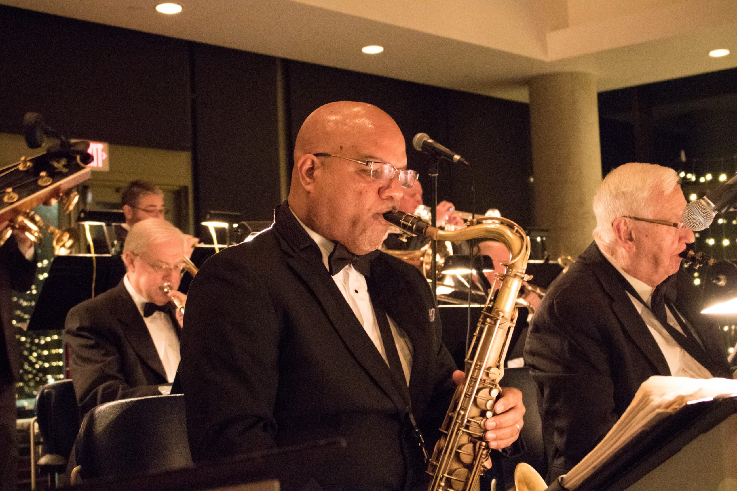 Bald Black man in a suit plays saxophone with white musicians around him.