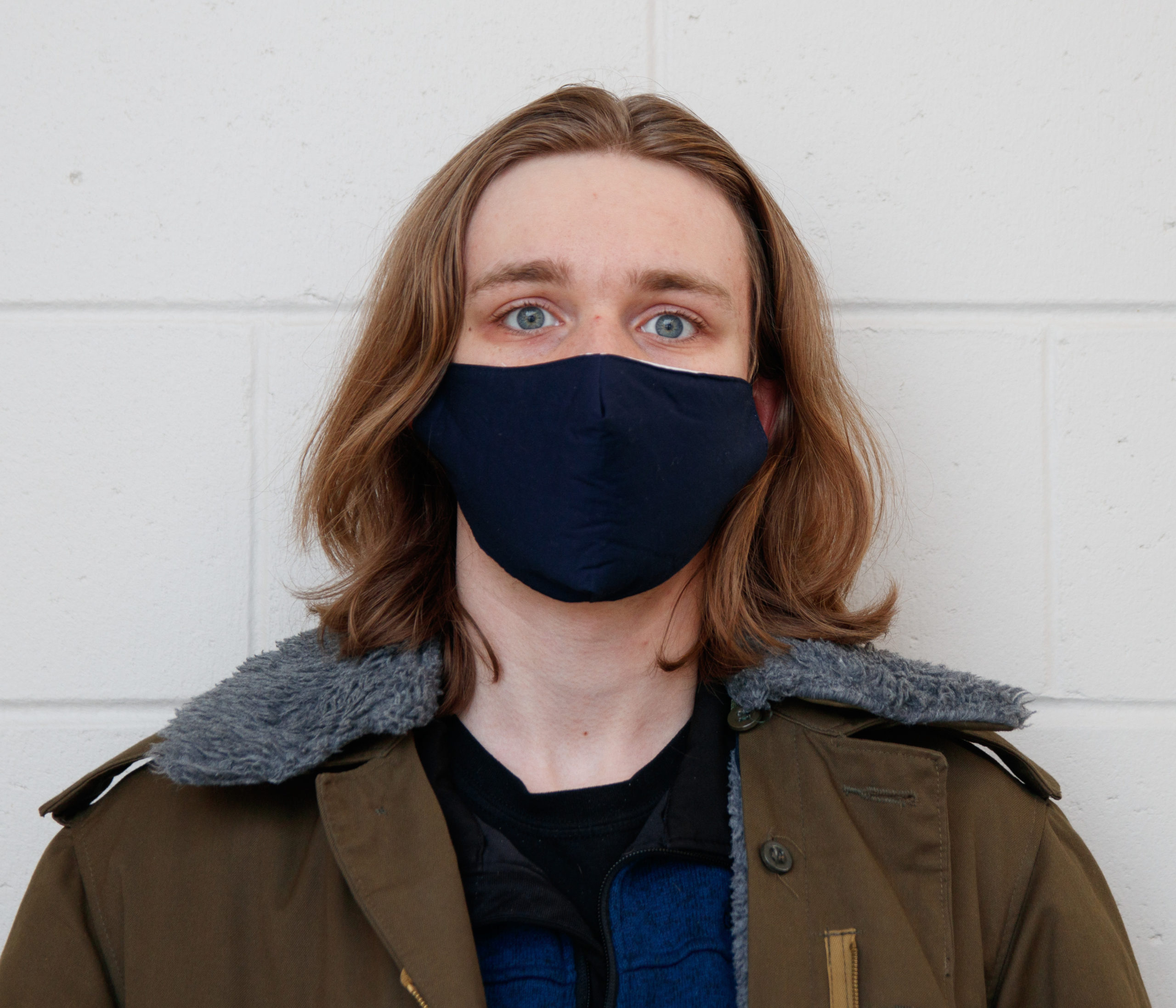 Male presenting person with shoulder length hair in mask wearing coat looks into camera on cement block background.