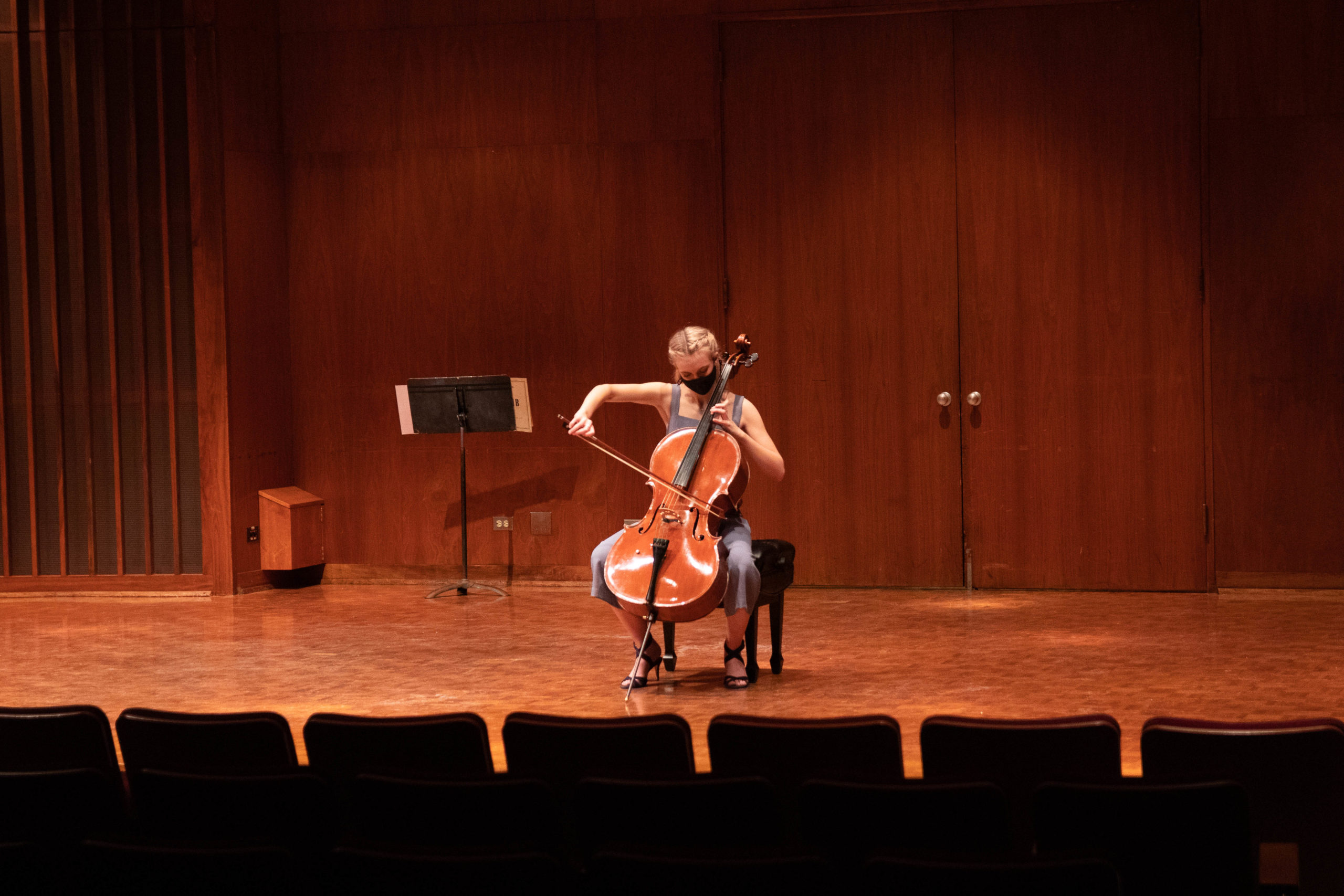 Masked young white person plays cello alone on stage.