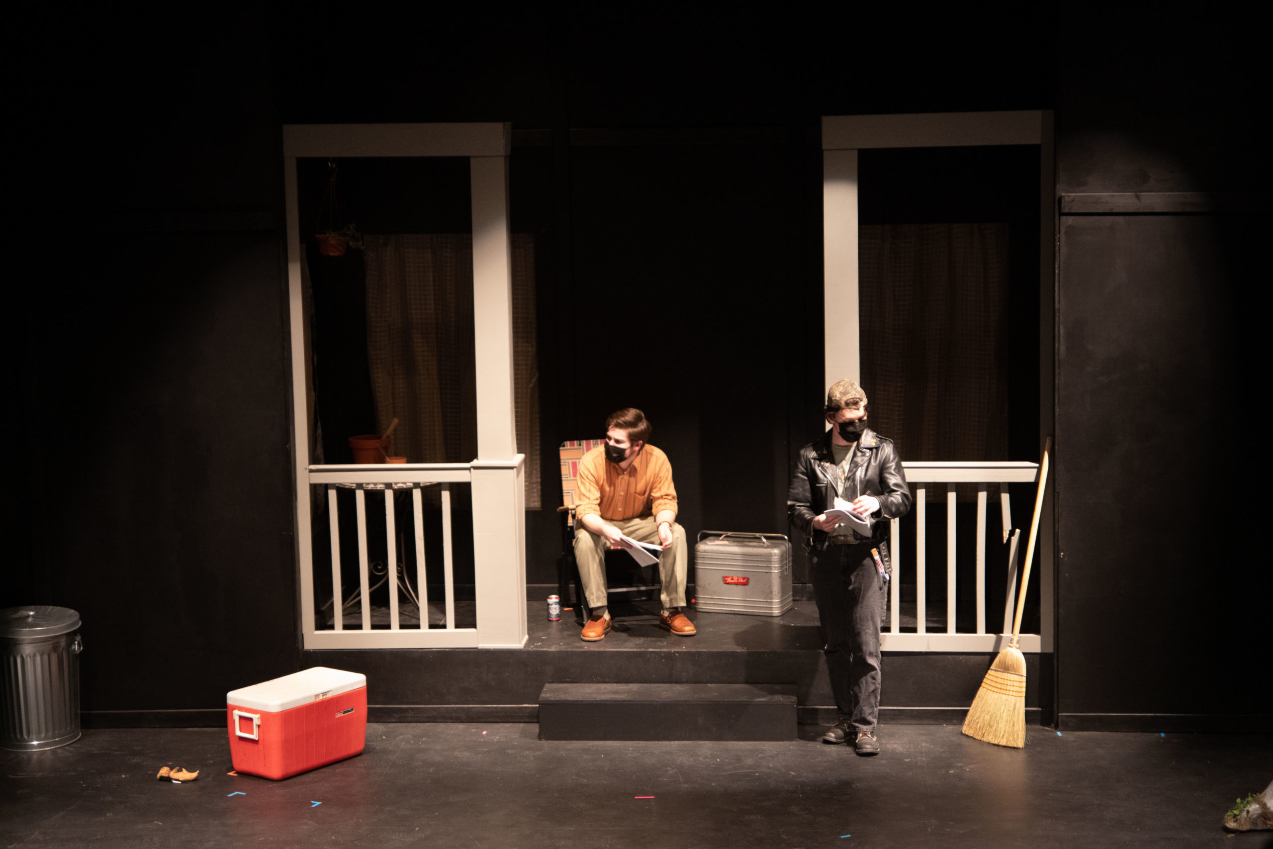 Black box stage with a house as the set, young white man sits on a chair on the porch and another young person stands near the steps