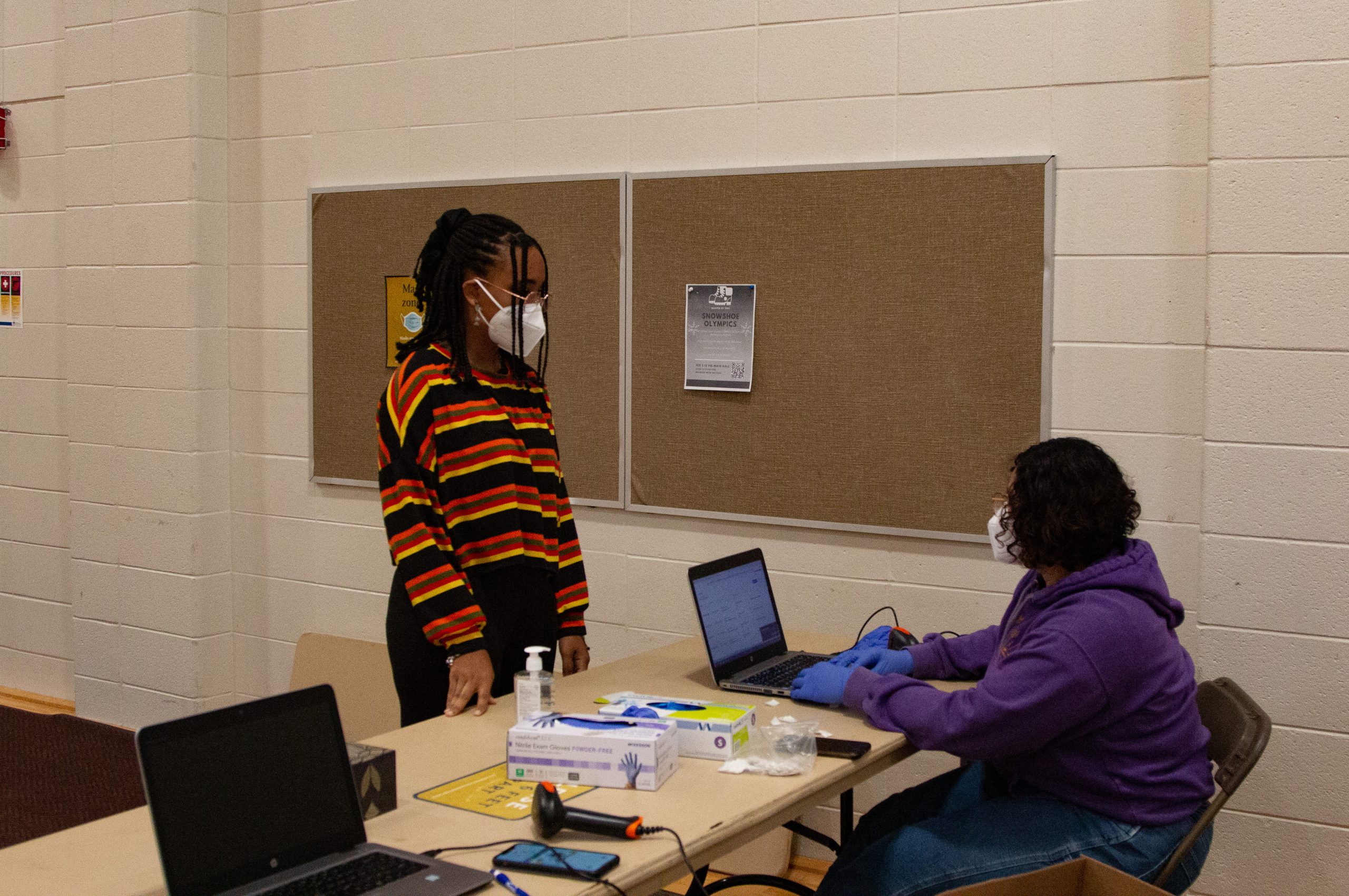 Person standing in mask talks to person wearing a mask sitting down at a computer.